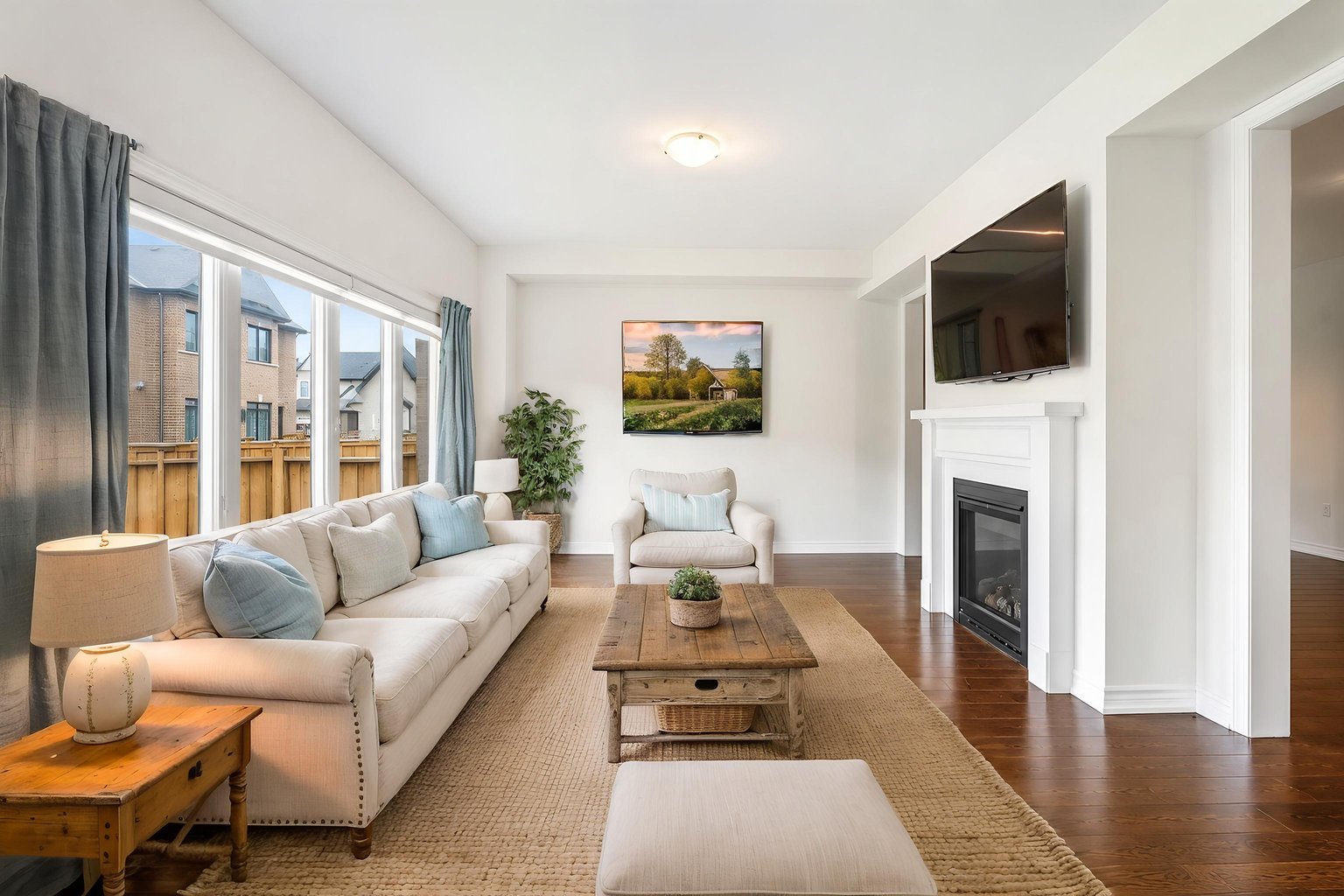 Coastal virtual staging of the same empty living room — white walls, dark hardwood, a fireplace with a white mantel and a wide side window — cream sofa with light-blue cushions, a reclaimed-wood coffee table, a jute rug and a landscape painting.